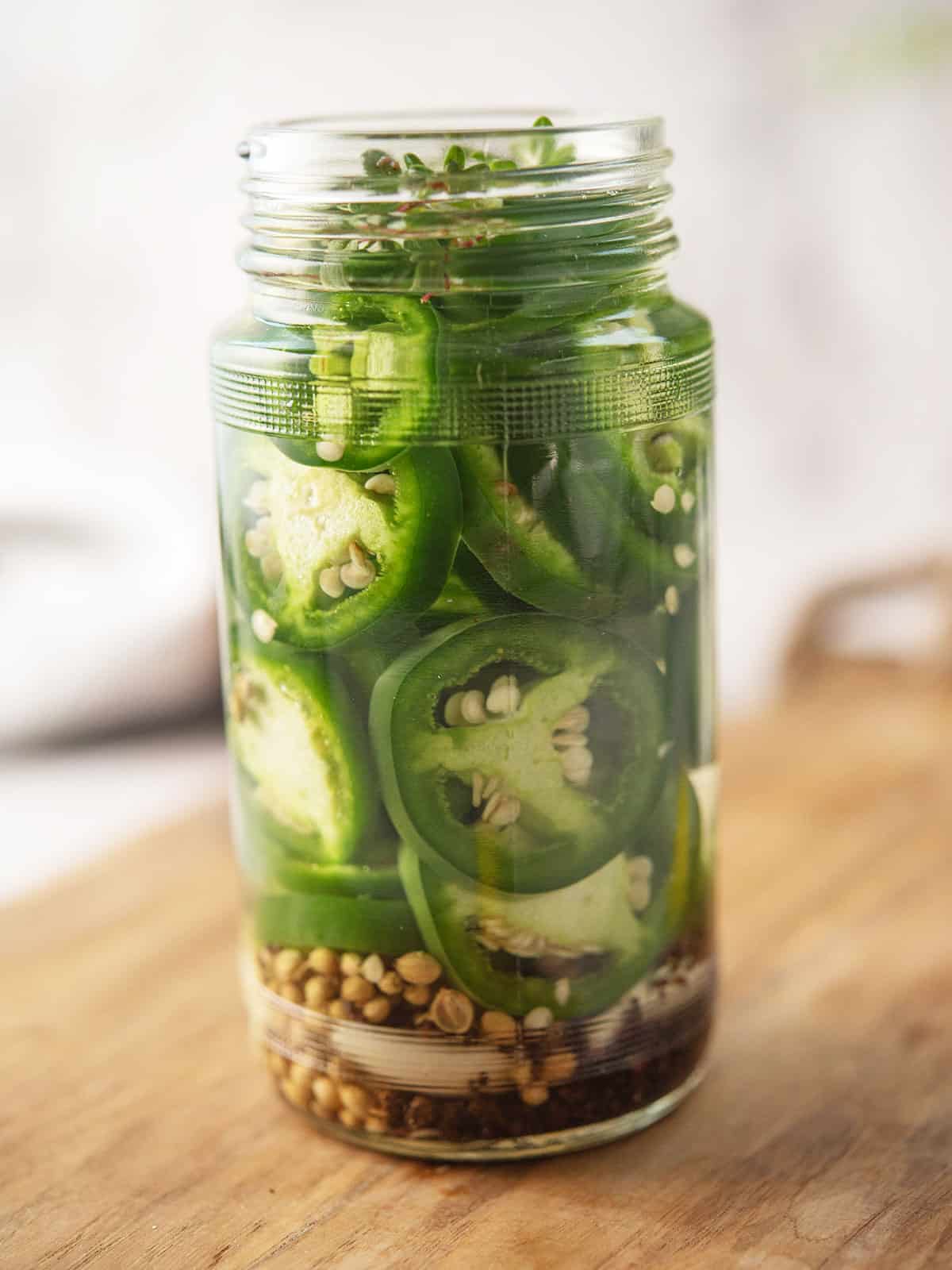 Jar of pickled jalapeños with fresh herbs on a wooden table.