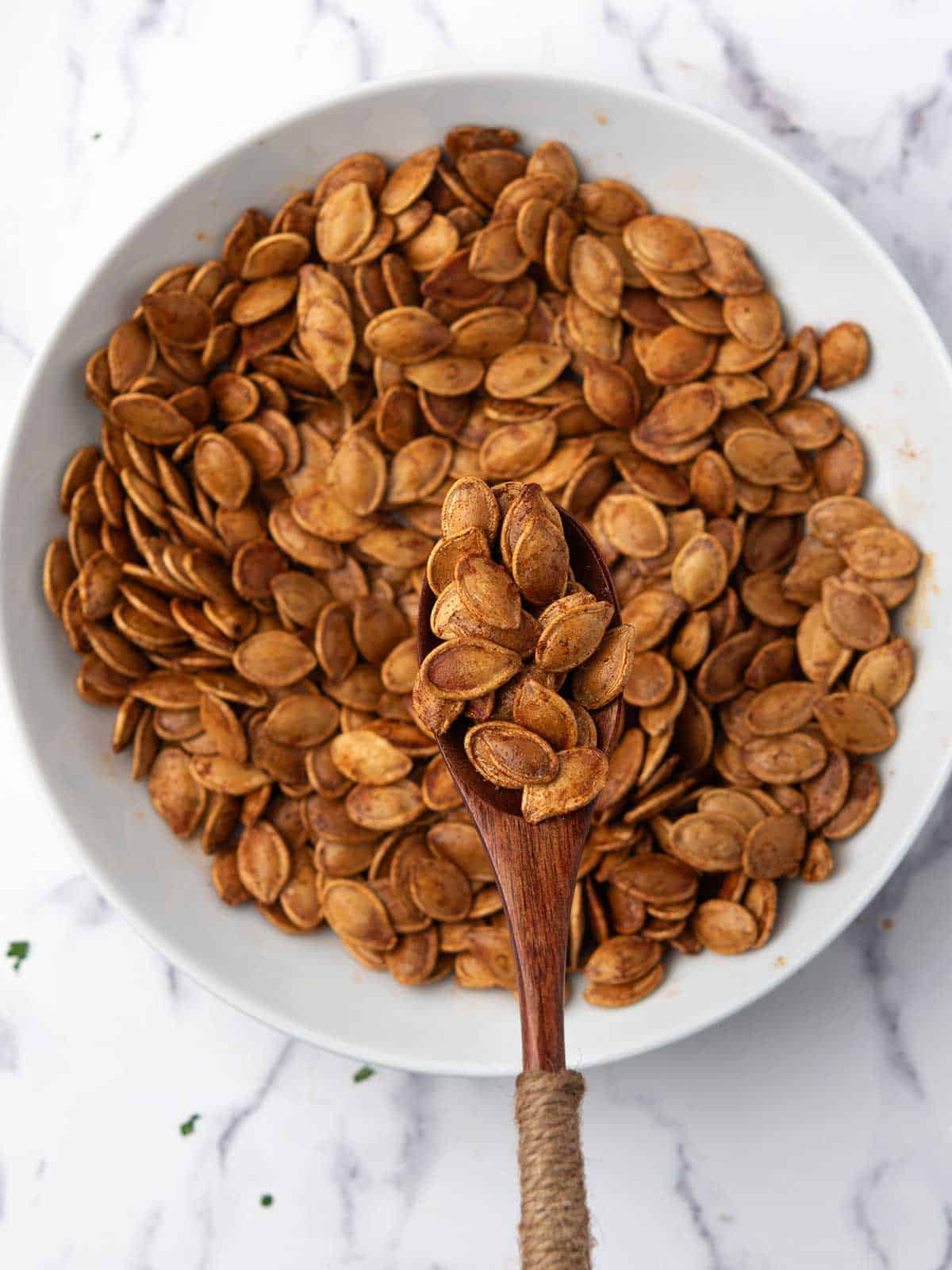 Bowl of golden brown spicy roasted pumpkin seeds with a wooden spoon holding a scoop.